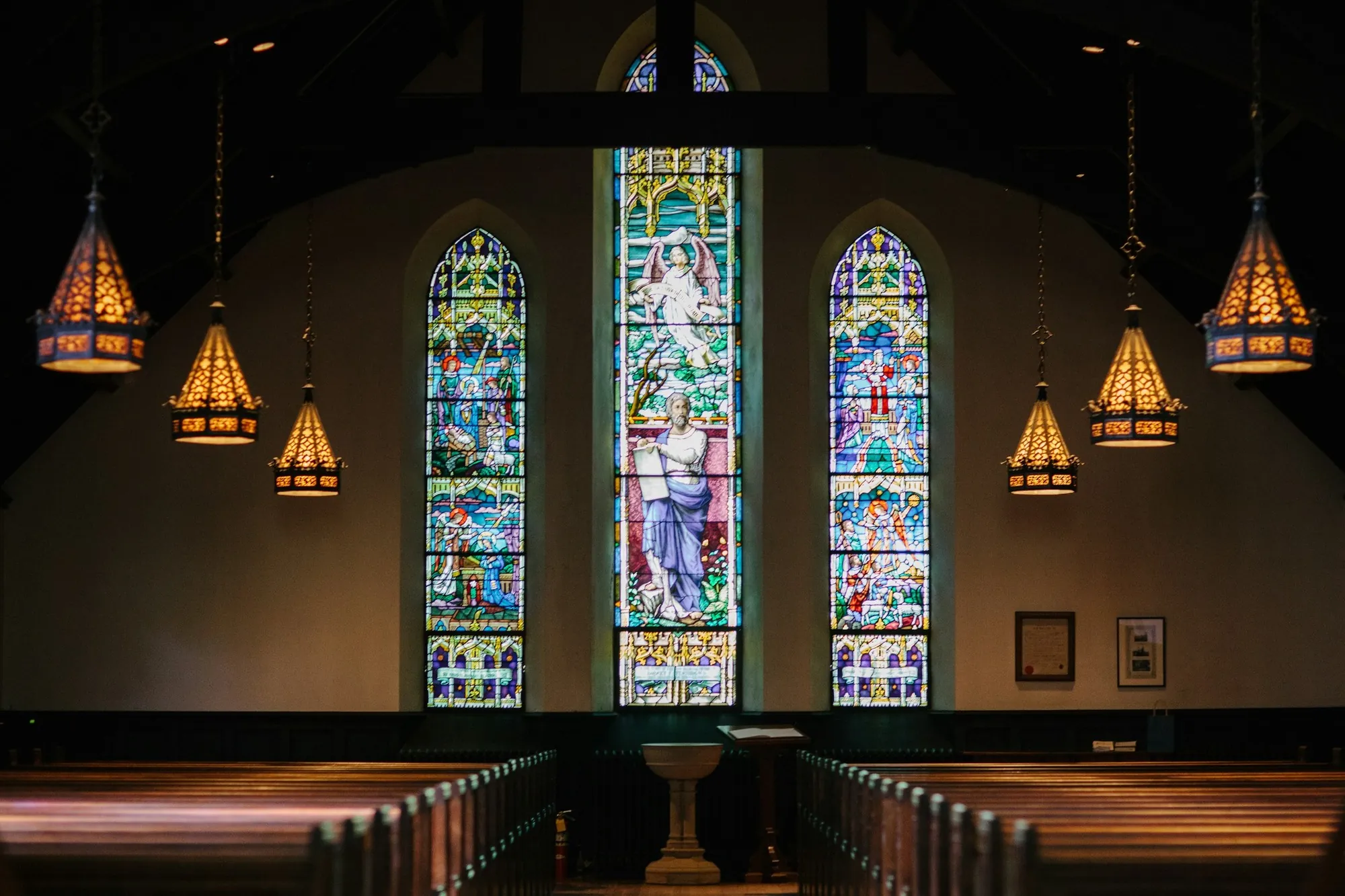 Inside a church: three stained-glass windows, hanging lamps and a bare wall on either side