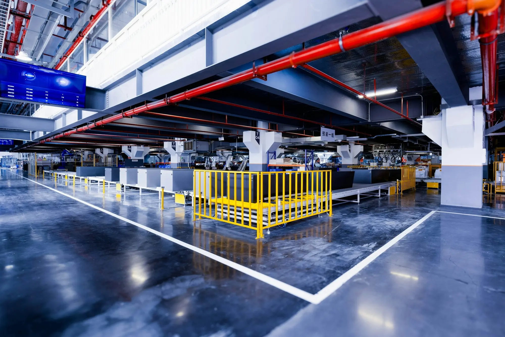 A production hall with conveyor lines, a yellow safety fence, a hanging display and a white column